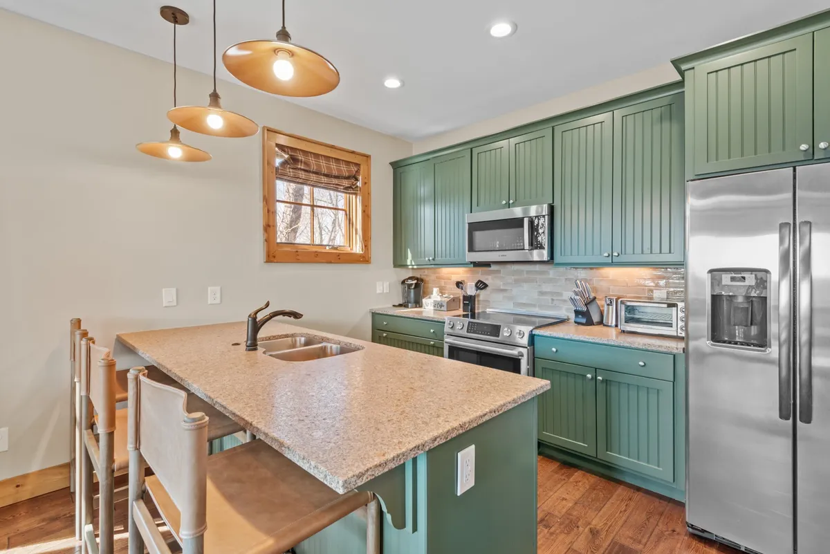A close-up view of the kitchen featuring green cabinetry, a granite countertop, pendant lighting, and a window with wooden blinds. The space feels both modern and inviting, emphasizing the cooking area and its readiness for family gatherings or casual meals.