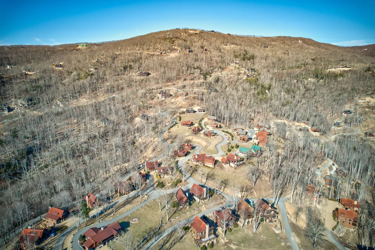 A wide exterior view with scattered cabins, winding roads, surrounding trees, and distant hills, set in a lush, green environment. The landscape appears serene, emphasizing the property's immersion in nature and the promise of seasonal beauty.