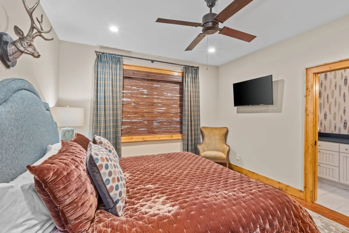 This image shows a king bedroom featuring a bed with plush bedding, decorative pillows, a chair, a window with blinds letting in natural light, and a wall-mounted TV. The cozy setup exudes comfort and is ideal for relaxation after a day of adventure.