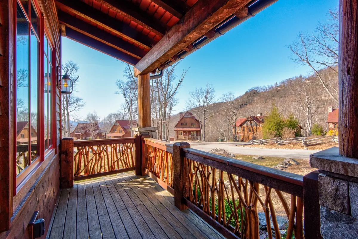 An exterior shot featuring a wooden porch railing with decorative patterns, neighboring houses nestled in the landscape, and a panoramic view of the mountains in the background. The perspective suggests a peaceful spot to relax and enjoy the outdoors.