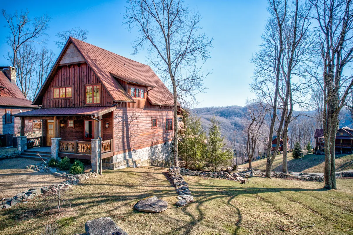 A picturesque view of a wooden cabin with a metal roof and stone foundation, featuring a spacious front porch overlooking serene mountain vistas. The scene evokes peace and intimacy, with the porch set up for morning coffee or evening stargazing, framed by the natural beauty of the surrounding landscape.