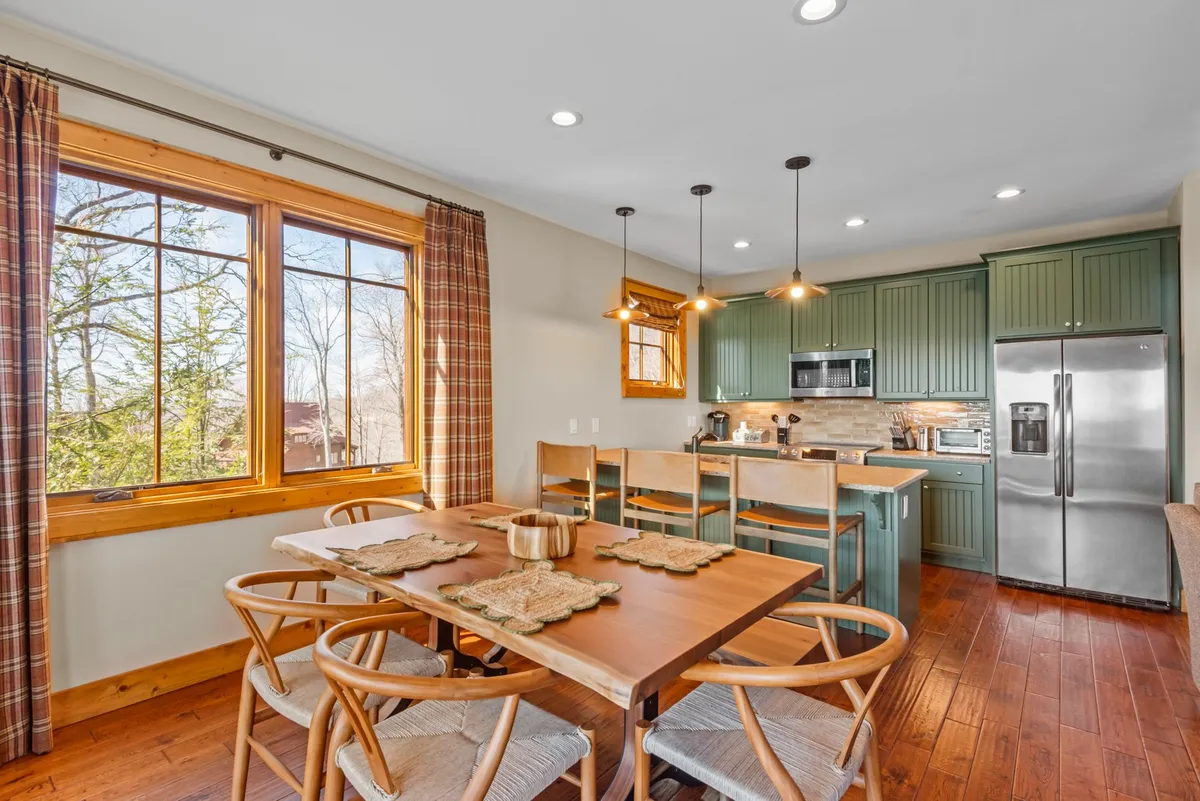 A spacious kitchen and dining area with a long wooden dining table set beside large windows, green cabinetry, and modern appliances. The table is ready for gatherings, while the open kitchen invites culinary activity. Soft, natural light fills the space, creating a warm and communal atmosphere.