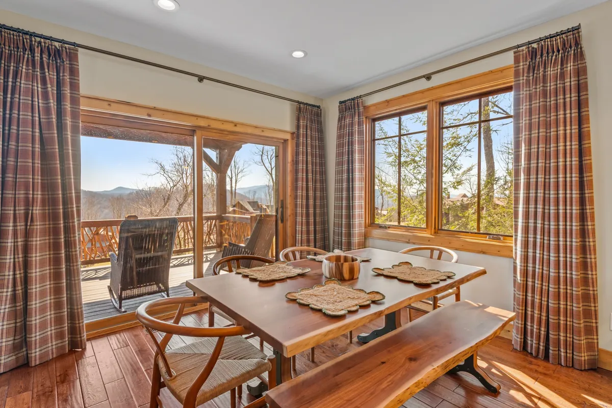 A cozy dining area with a wooden table surrounded by windows dressed with curtains, wooden flooring, and soft lighting. The intimate atmosphere invites families and friends to gather for meals, conversations, or game nights, with mountain views just beyond the glass.