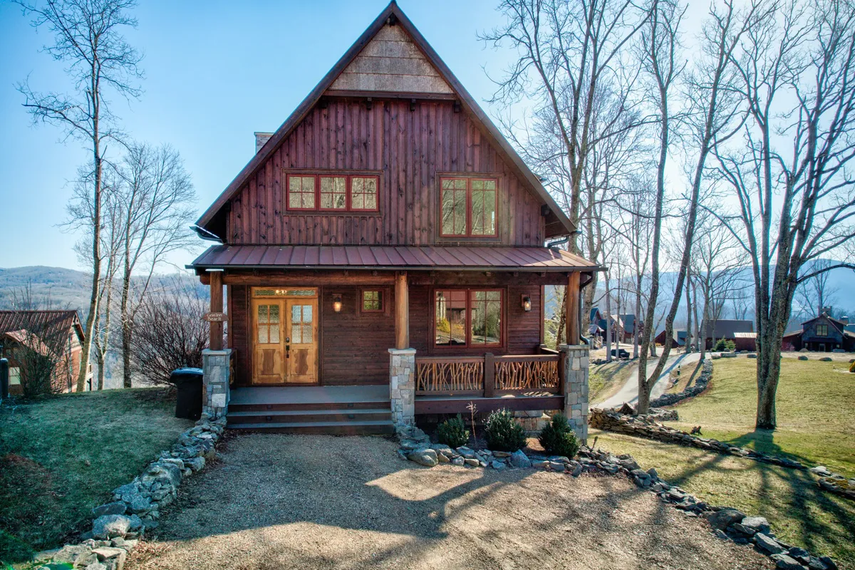The image showcases the inviting exterior of a charming mountain cabin with a wooden facade, sturdy stone pillars, and a welcoming front porch. The scene suggests a cozy mountain escape perfect for gathering, with the porch offering a prime spot to relax and take in sweeping views or sunsets.