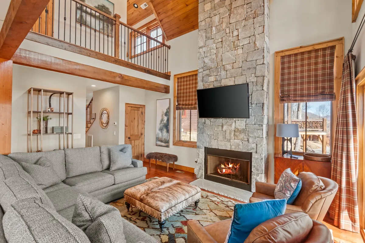 The great room with a large sectional sofa centered around a grand stone fireplace, exposed wooden beams overhead, mountain-themed artwork, and floor-to-ceiling windows that reveal the stunning landscape beyond. The inviting layout encourages gatherings and relaxation after a day of adventure.