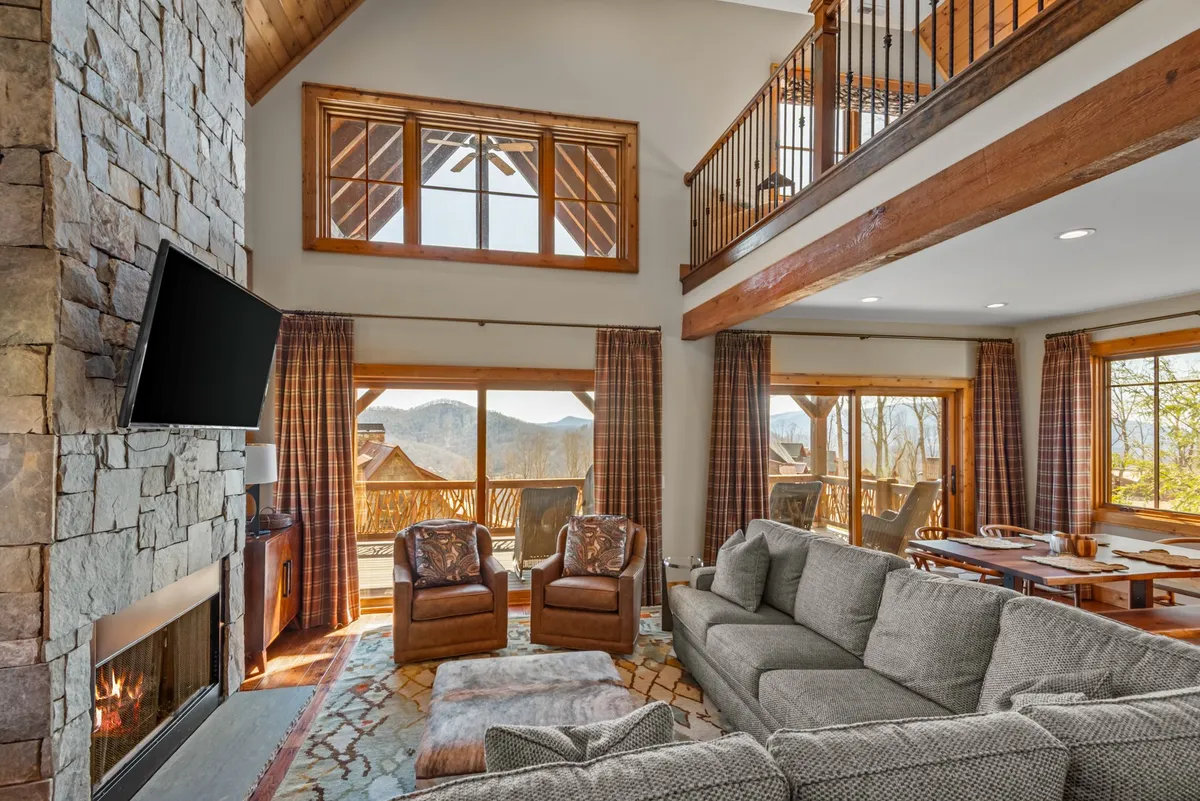 An open-concept living and dining area where a stone fireplace anchors the space. The seating area is cozy and designed for both relaxation and socializing, while a dining table is within easy reach, reflecting seamless flow between lounging and dining. Large windows pull in natural light and offer glimpses of mountain scenery.