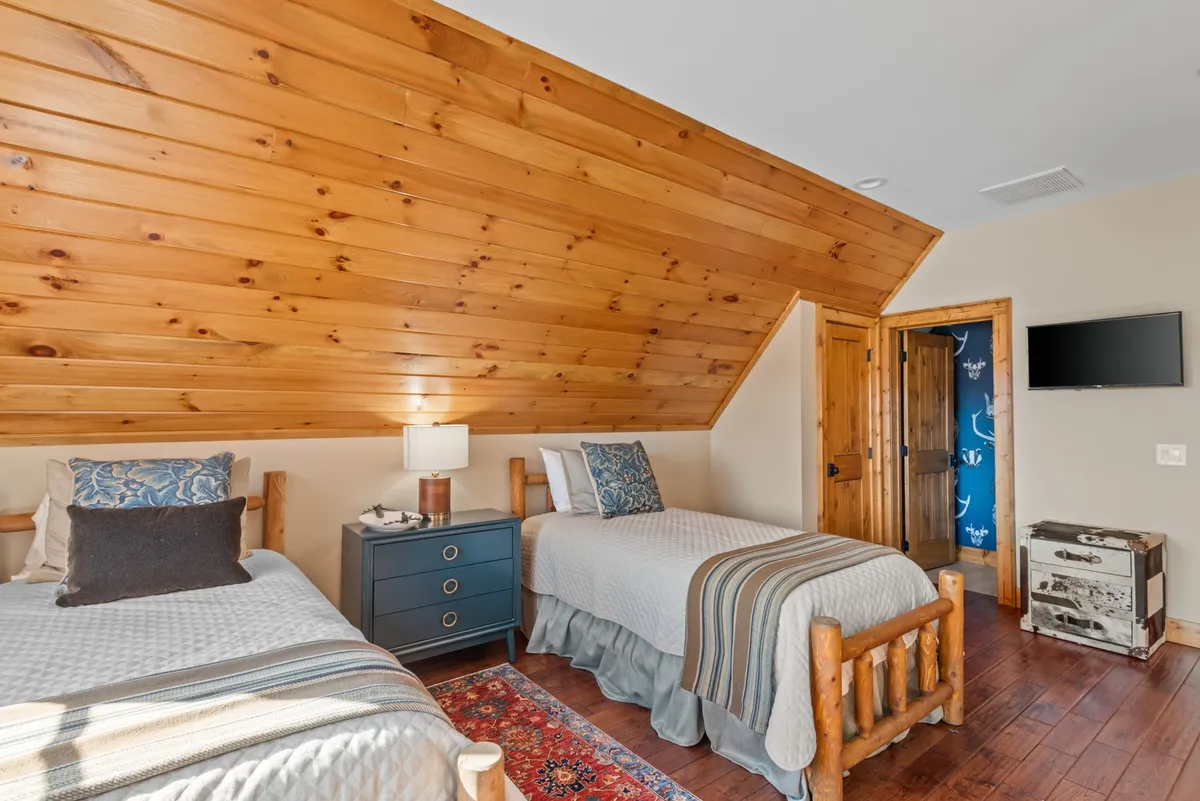 A charming loft space featuring two twin beds with decorative pillows, a nightstand with a lamp, a rug, and a wooden ceiling. The area feels whimsical and perfect for children or playful sleepovers.