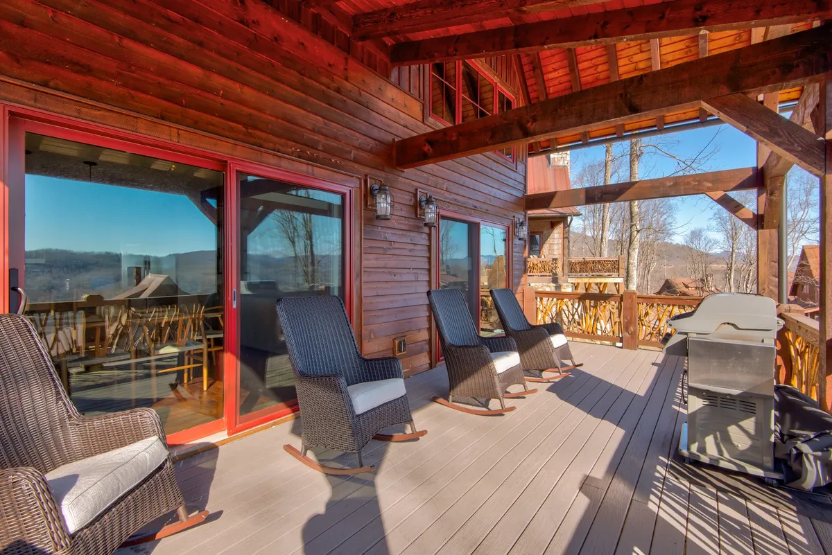 An exterior shot showcasing deck chairs and a grill set against large windows and wooden beams, with scenic views perfect for relaxing, grilling, and socializing in a tranquil mountain setting.