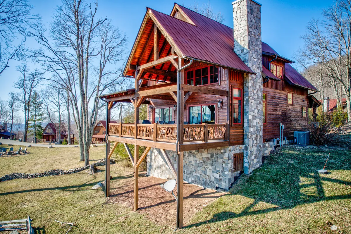 This exterior image features a spacious wooden deck with a stone chimney, a distinctive red metal roof, and large windows overlooking the surrounding natural landscape. The setting is perfect for outdoor gatherings, barbecues, and enjoying the sunset.