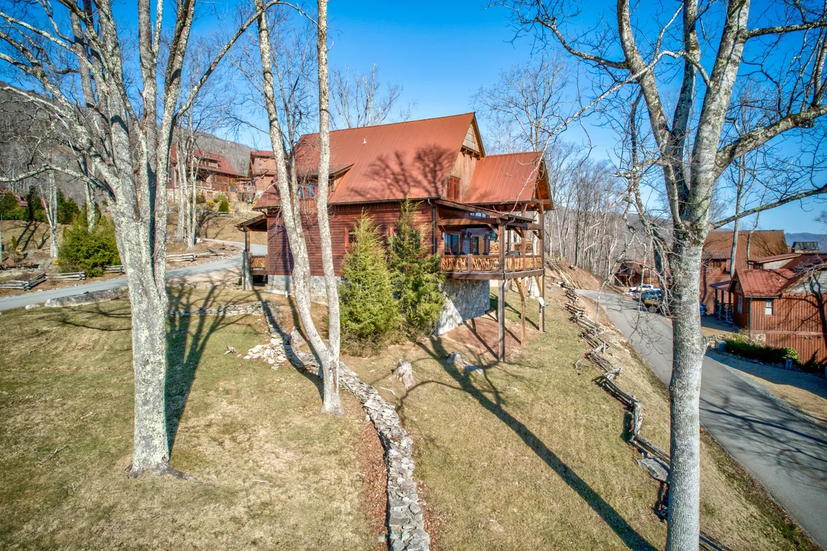 A charming rustic wooden cabin with a stone foundation sits nestled among mature trees and landscaped grounds. The image captures the inviting front porch at sunset, where comfortable seating beckons guests to relax, enjoy a glass of wine, or revel in the tranquil mountain setting. The surrounding natural elements and warm glow create a private and romantic atmosphere.