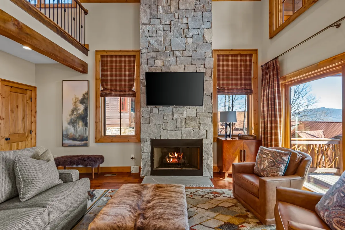 A warmly lit great room featuring a prominent stone fireplace flanked by large windows, comfortable seating arranged for conversation, and playful artwork that hints at mountain themes. The windows let in abundant sunlight and frame a view of the mountain landscape outside, enhancing the welcoming and cozy atmosphere of the room.